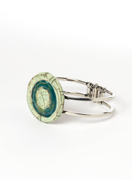 Bracciale rigido in metallo con elemento centrale in ceramica Raku smaltata, caratterizzato da un disco concentrico che richiama le orbite lunari. I toni verde chiaro e ottanio si fondono tra loro con delicate venature e crepe tipiche della tecnica Raku
