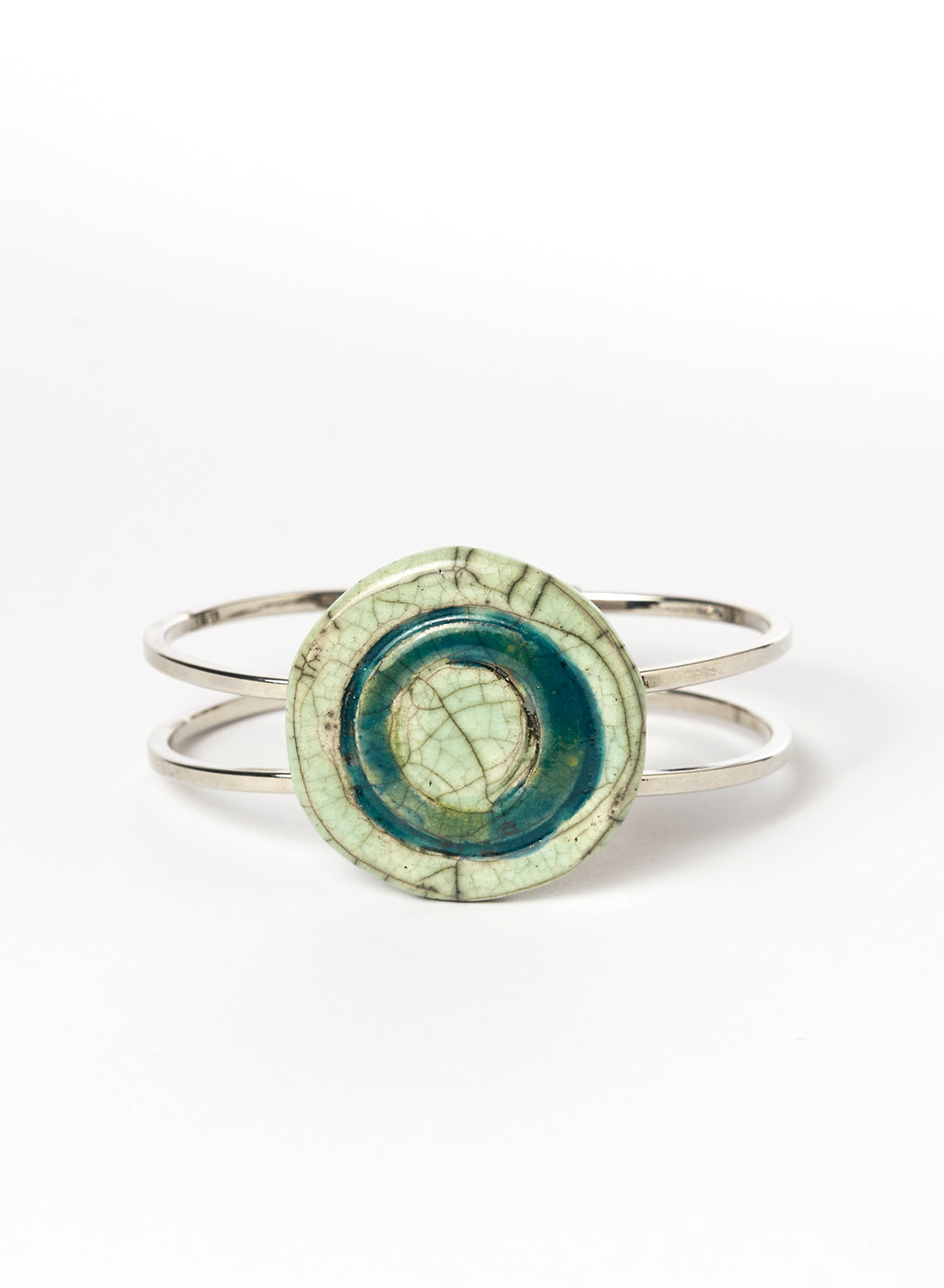 Bracciale rigido in metallo con elemento centrale in ceramica Raku smaltata, caratterizzato da un disco concentrico che richiama le orbite lunari. I toni verde chiaro e ottanio si fondono tra loro con delicate venature e crepe tipiche della tecnica Raku