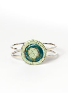 Bracciale rigido in metallo con elemento centrale in ceramica Raku smaltata, caratterizzato da un disco concentrico che richiama le orbite lunari. I toni verde chiaro e ottanio si fondono tra loro con delicate venature e crepe tipiche della tecnica Raku