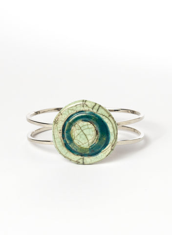 Bracciale rigido in metallo con elemento centrale in ceramica Raku smaltata, caratterizzato da un disco concentrico che richiama le orbite lunari. I toni verde chiaro e ottanio si fondono tra loro con delicate venature e crepe tipiche della tecnica Raku