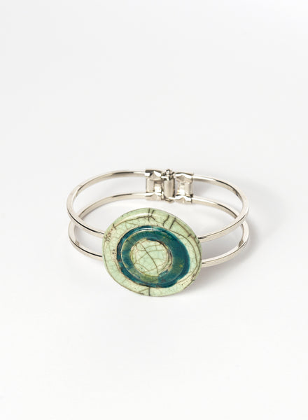 Bracciale rigido in metallo con elemento centrale in ceramica Raku smaltata, caratterizzato da un disco concentrico che richiama le orbite lunari. I toni verde chiaro e ottanio si fondono tra loro con delicate venature e crepe tipiche della tecnica Raku