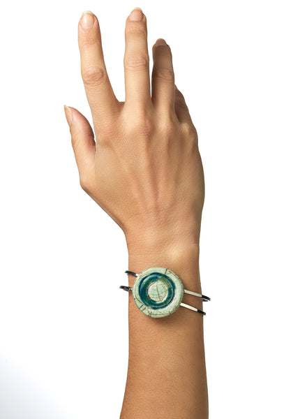 Bracciale rigido in metallo con elemento centrale in ceramica Raku smaltata, caratterizzato da un disco concentrico che richiama le orbite lunari. I toni verde chiaro e ottanio si fondono tra loro con delicate venature e crepe tipiche della tecnica Raku