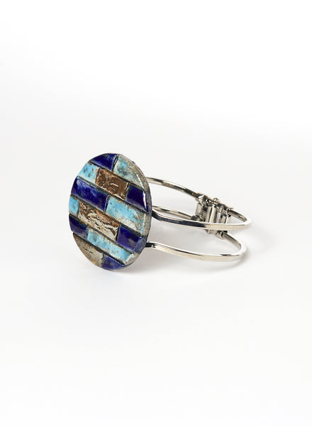 Bracciale rigido in metallo lucido con elemento in ceramica Raku, caratterizzato da un disco decorato con inseriti a rilievo in tonalità marine: blu, turchese e oro. 