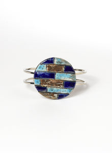 Bracciale rigido in metallo lucido con elemento in ceramica Raku, caratterizzato da un disco decorato con inseriti a rilievo in tonalità marine: blu, turchese e oro. 