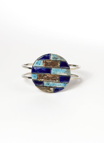 Bracciale rigido in metallo lucido con elemento in ceramica Raku, caratterizzato da un disco decorato con inseriti a rilievo in tonalità marine: blu, turchese e oro. 