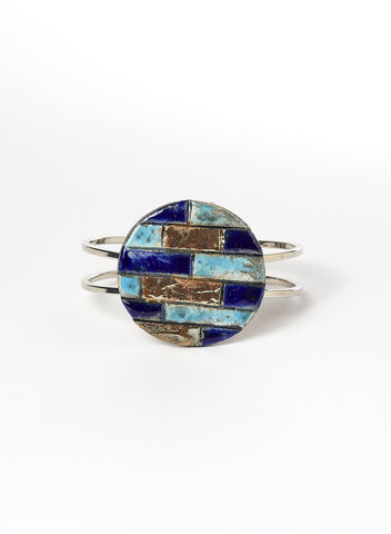 Bracciale rigido in metallo lucido con elemento in ceramica Raku, caratterizzato da un disco decorato con inseriti a rilievo in tonalità marine: blu, turchese e oro. 