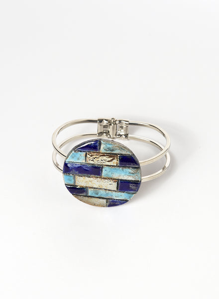 Bracciale rigido in metallo lucido con elemento in ceramica Raku, caratterizzato da un disco decorato con inseriti a rilievo in tonalità marine: blu, turchese e oro. 
