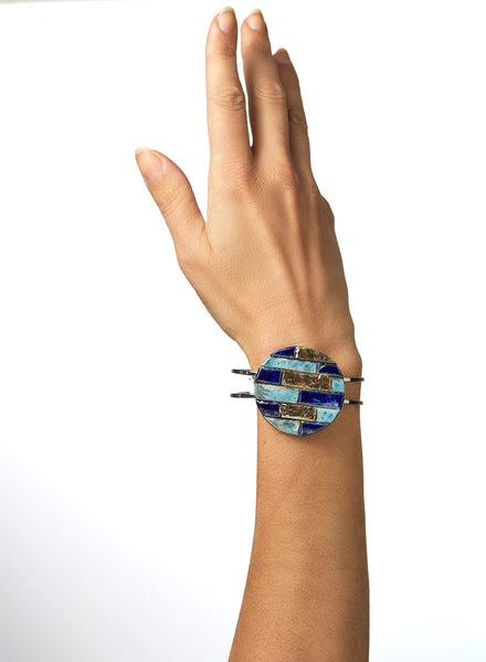 Bracciale rigido in metallo lucido con elemento in ceramica Raku, caratterizzato da un disco decorato con inseriti a rilievo in tonalità marine: blu, turchese e oro. 